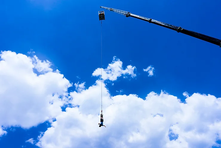 A man suspended from a crane, with a clear sky and scattered clouds in the background.