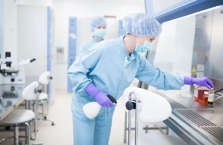 A lab assistant in a blue uniform, wearing a mask and cap, sanitizes equipment in a laboratory