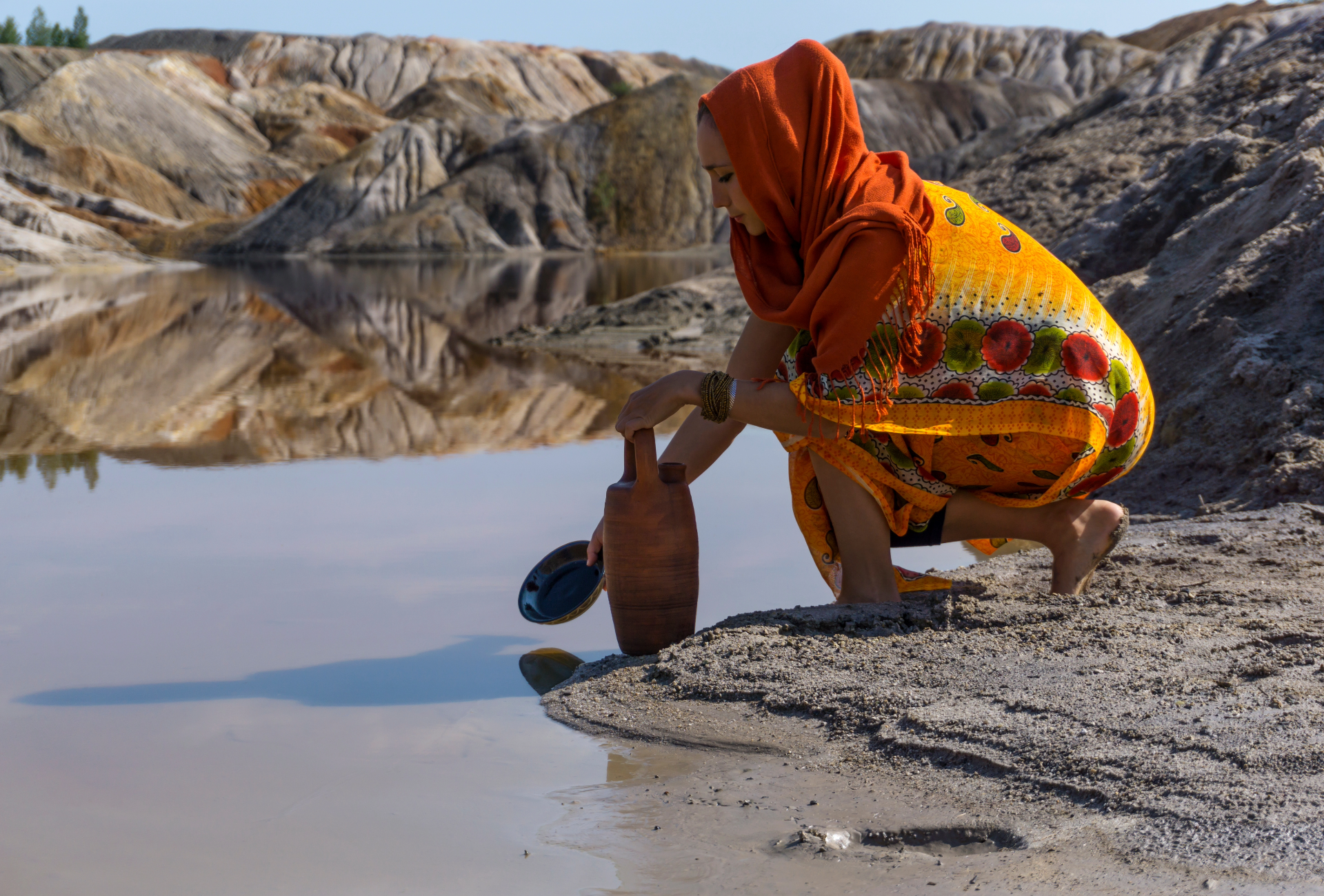 a women sitting next to a river filling water in a wooden bottle
