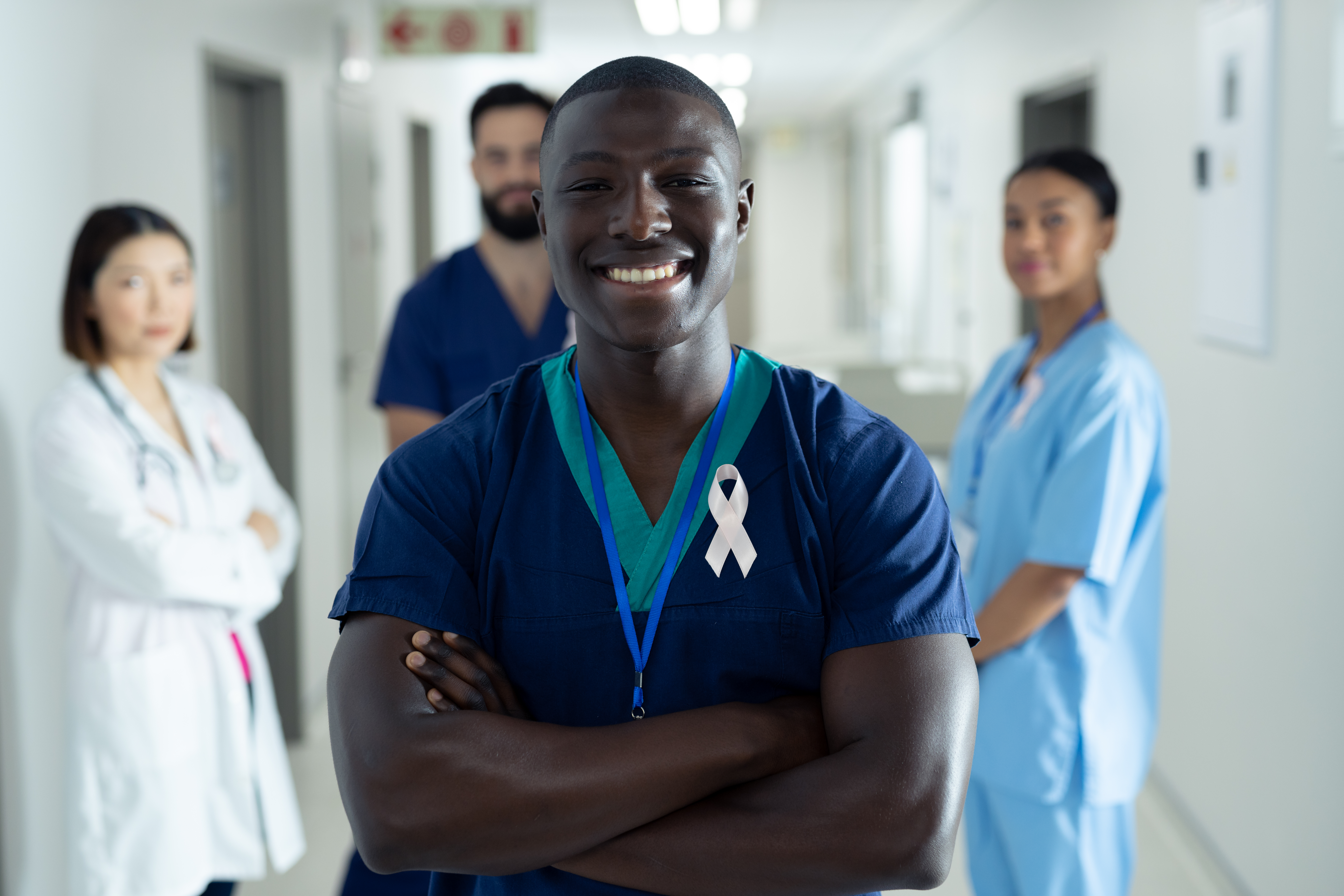 Portrait of happy african american male healthcare worker with cancer ribbon in hospital corridor. Hospital, medical and healthcare services, copy space.